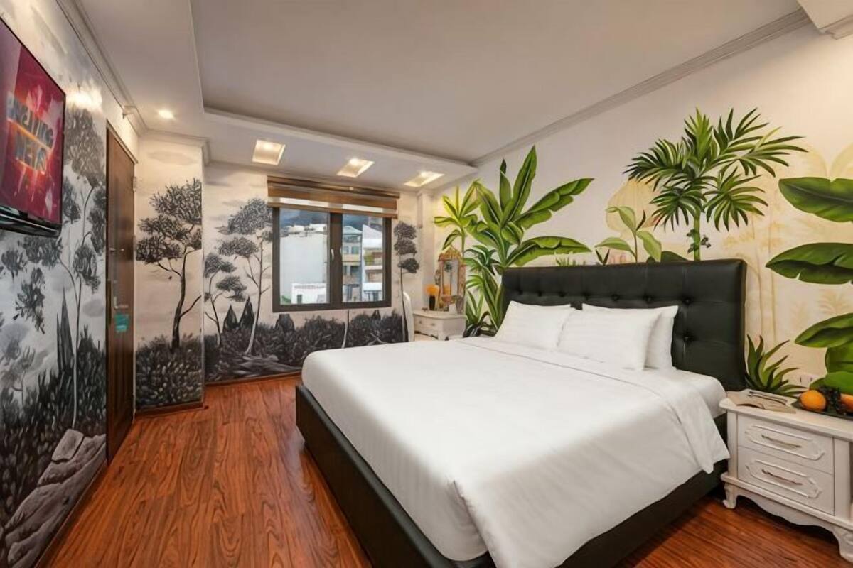 Un dormitorio con una cama grande, una mesita de noche, un televisor montado en la pared y una ventana con vistas.