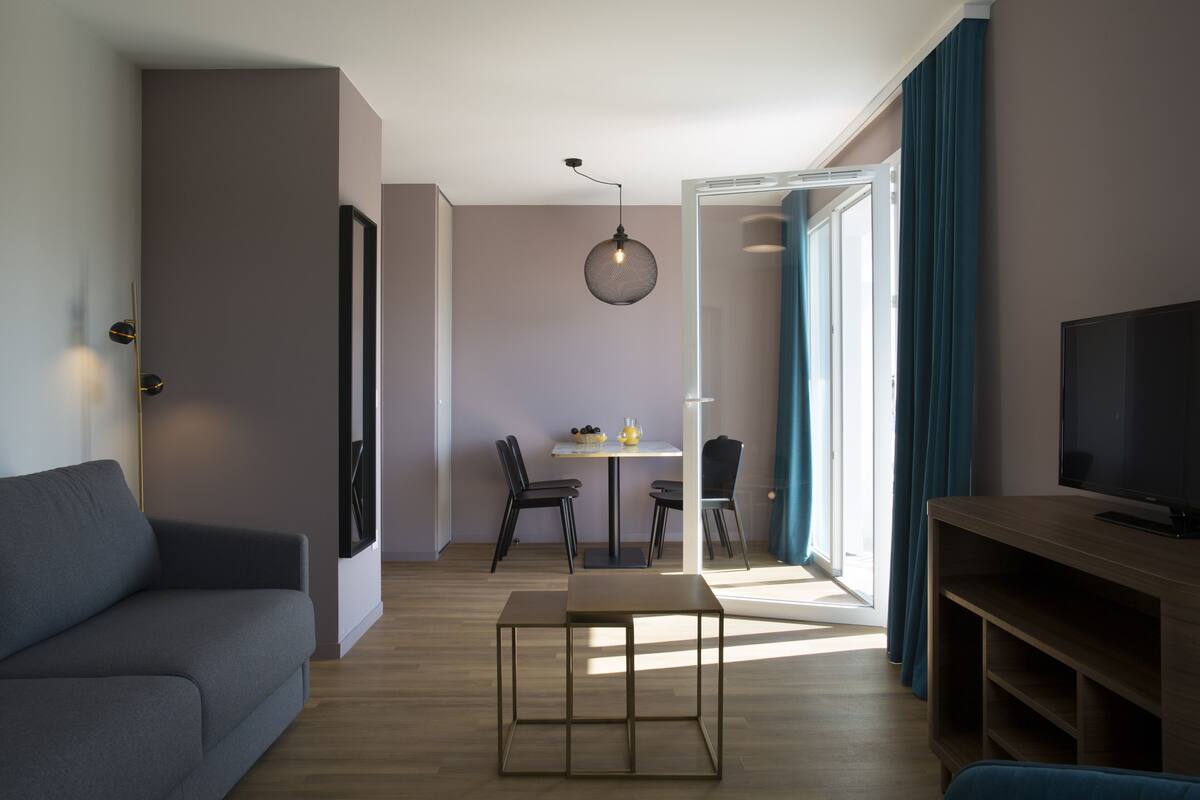 Un salon moderne comprenant un canapé, un meuble pour la télévision, une table à manger avec des chaises et une grande fenêtre avec des rideaux.