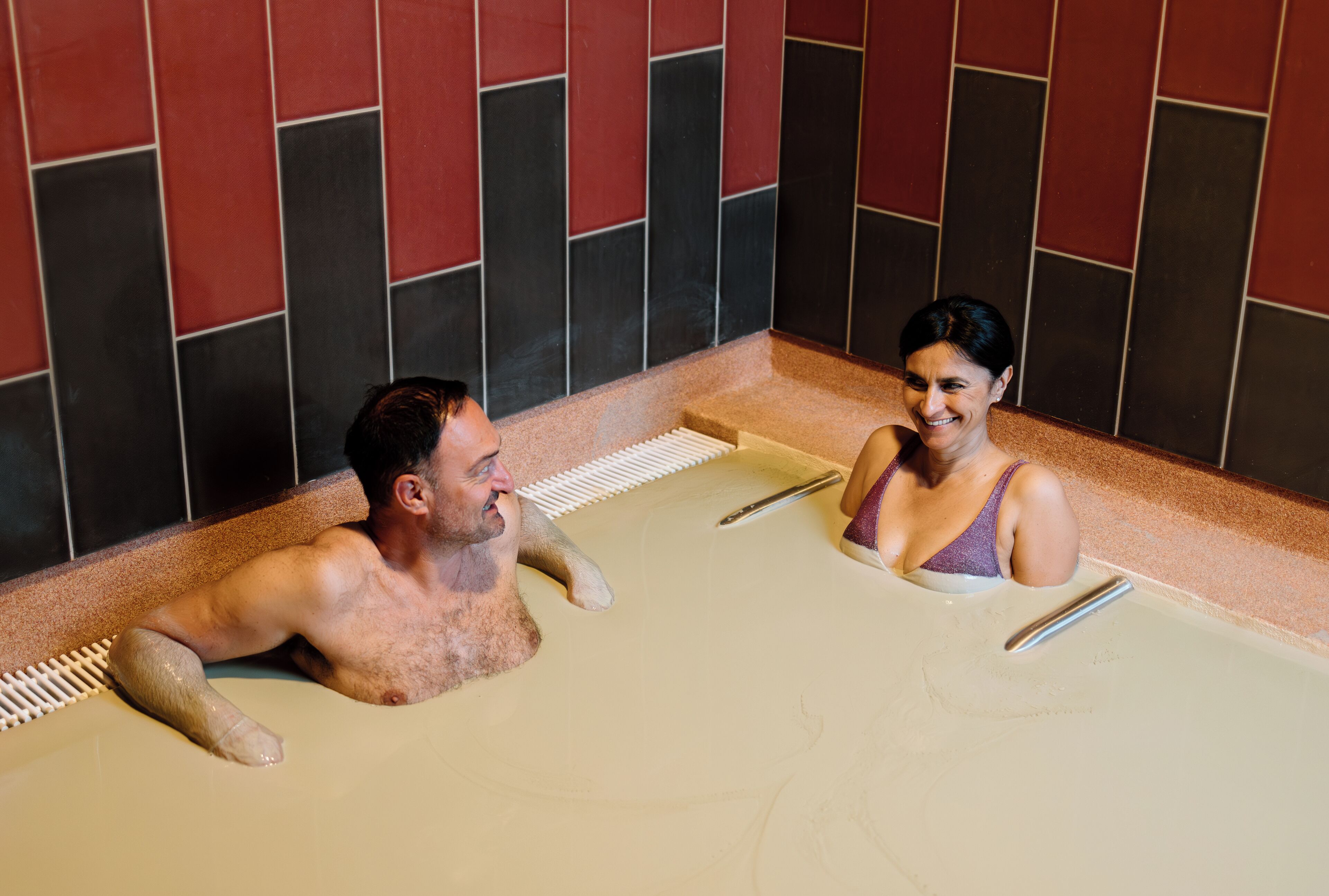 A man and a woman in a bathtub with a tiled wall in the background.