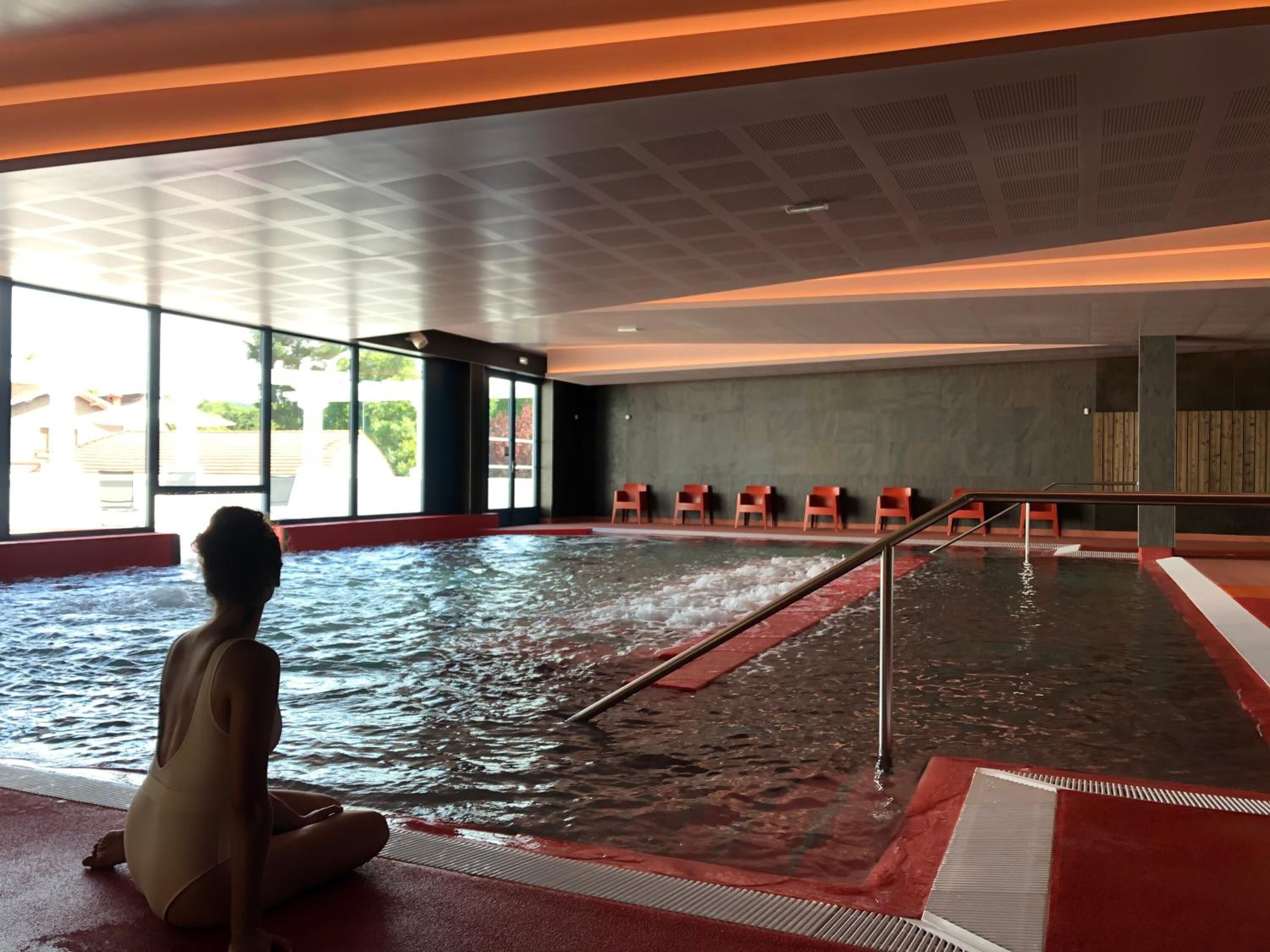 An indoor swimming pool with a person sitting by the edge, large windows, and a row of chairs along the wall.