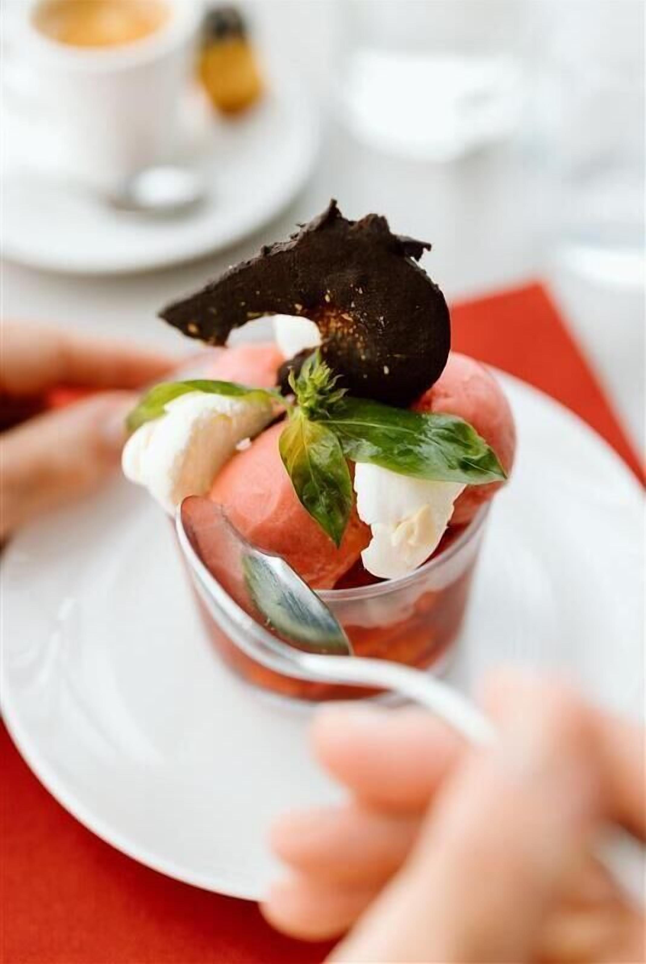 Postre con una bola de helado, un trozo de chocolate y una hoja de albahaca sobre un plato blanco.