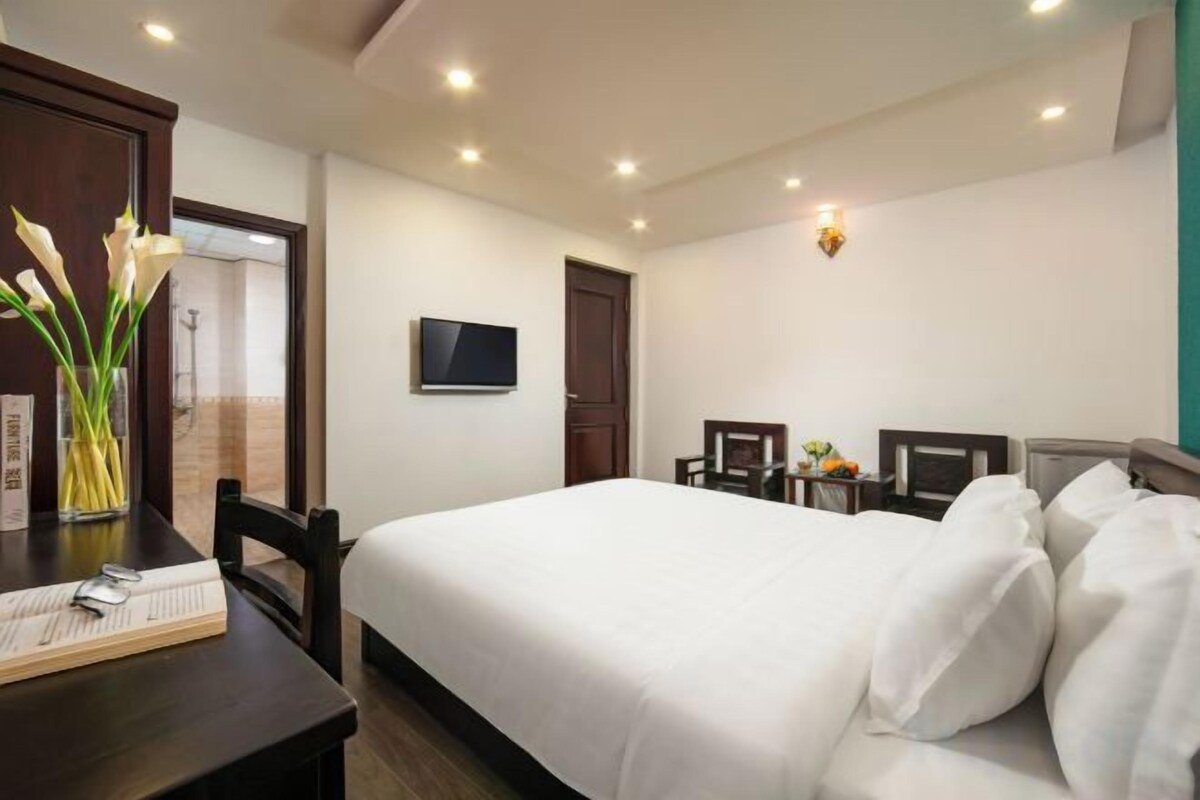 A hotel room with a bed, a desk with a vase of flowers, a television on the wall, and a door leading to a bathroom.