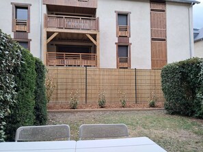 A building with a wooden balcony, a fenced yard, and a table with chairs.