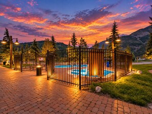 Une piscine extérieure entourée d’une clôture, avec un chemin en briques, bordée d’arbres et offrant une vue sur un paysage montagneux.