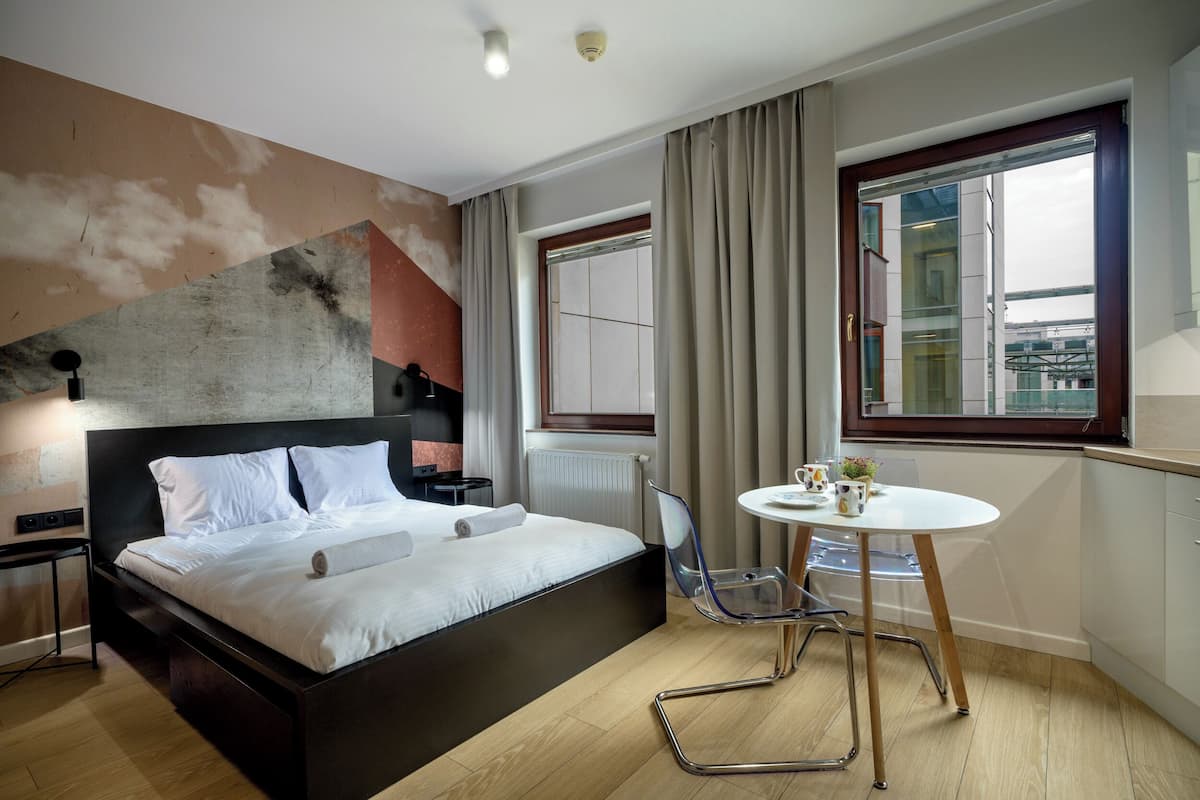 A modern hotel room with a large bed, a small round table with a chair, a window with curtains, and a view of the city.