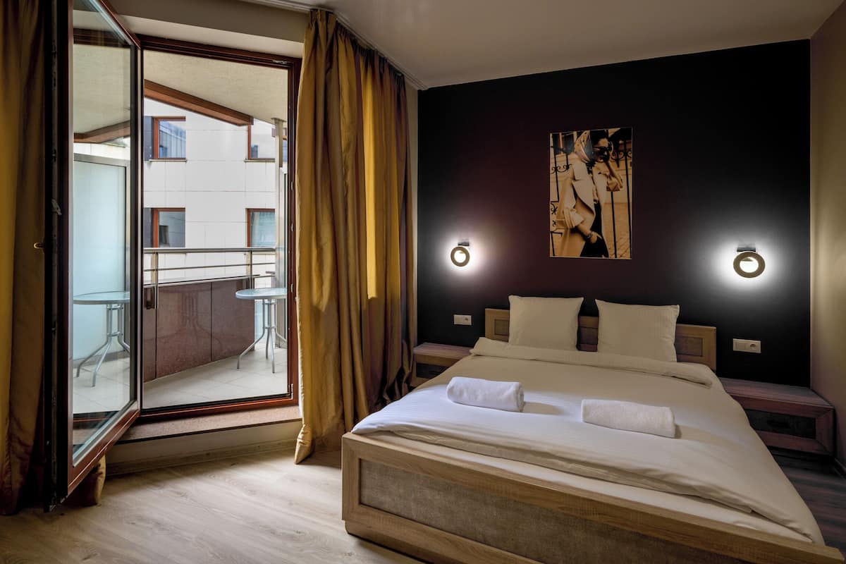 A hotel room with a large bed, a balcony with a view of buildings, and two wall-mounted lights.