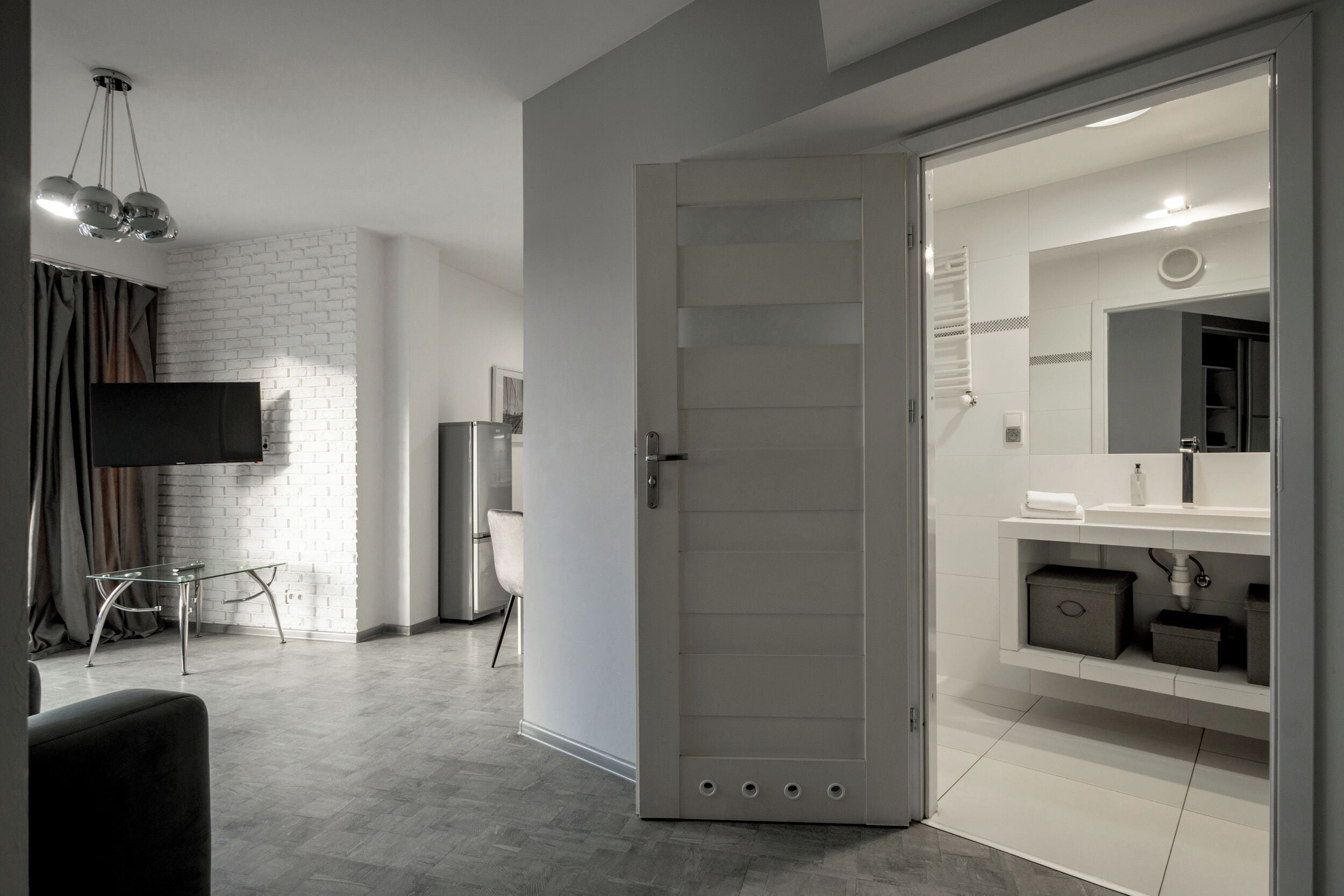 A modern room with a white brick wall, a flat-screen TV mounted on the wall, a glass table, a chair, a wardrobe with a mirror, and a built-in sink.