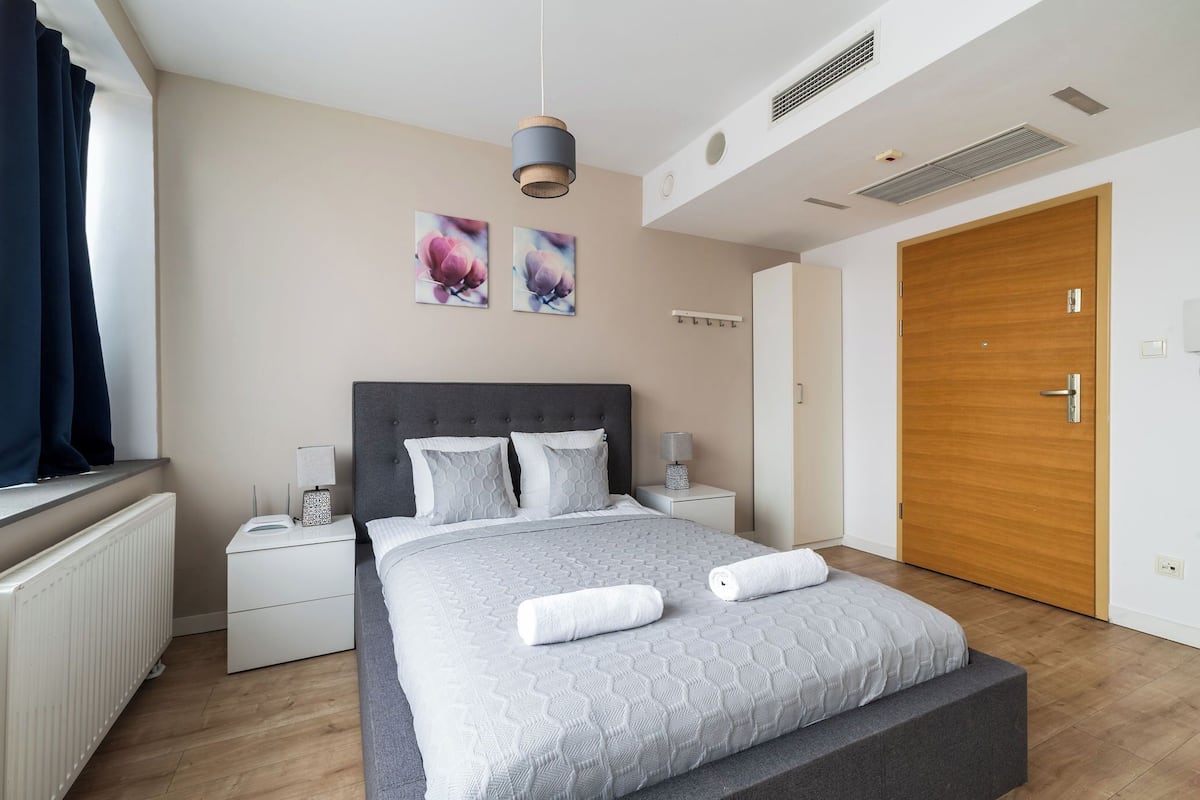 A modern bedroom with a large bed, bedside tables, a wooden door, and a window with curtains.