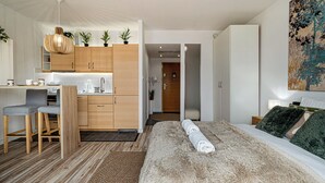 Ein modernes Hotelzimmer mit einem großen Bett, einer Sitzecke mit Schreibtisch und Stühlen, einer Küchenzeile und einem durch eine offene Tür einsehbaren Badezimmer.