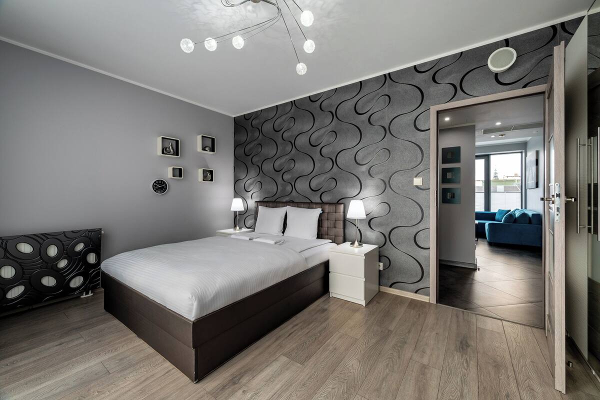 A modern bedroom with a large bed, bedside tables with lamps, a patterned wall, and a view into another room.