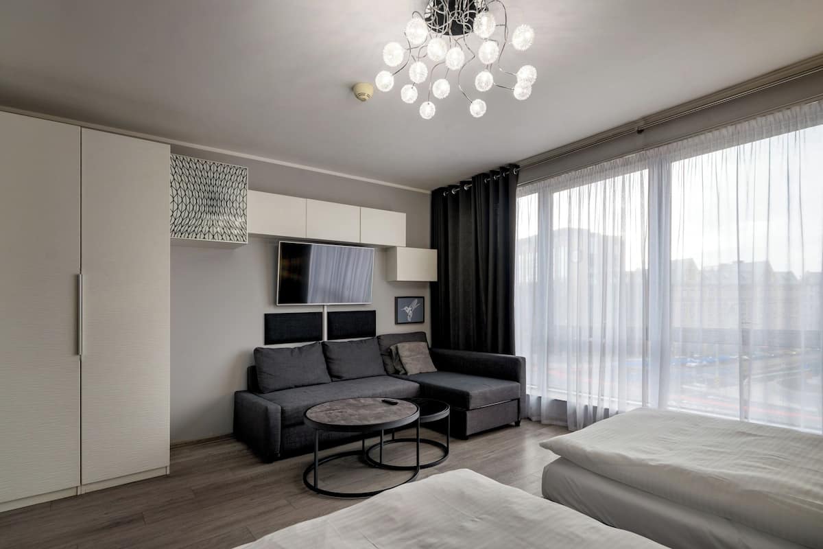 A modern hotel room with a grey sofa, a round coffee table, a bed, and a large window with sheer curtains.