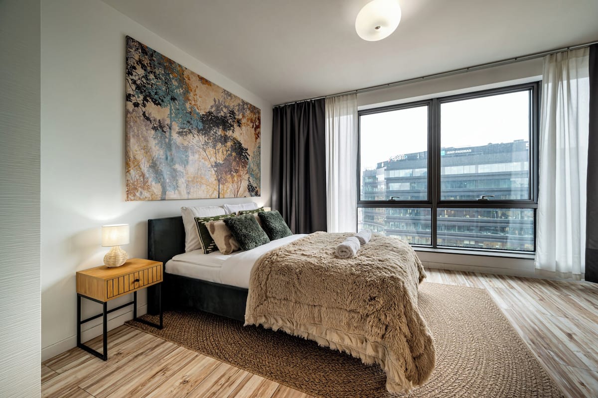 A modern bedroom with a large window, a bed with a fluffy blanket, a side table with a lamp, and a large abstract painting on the wall.