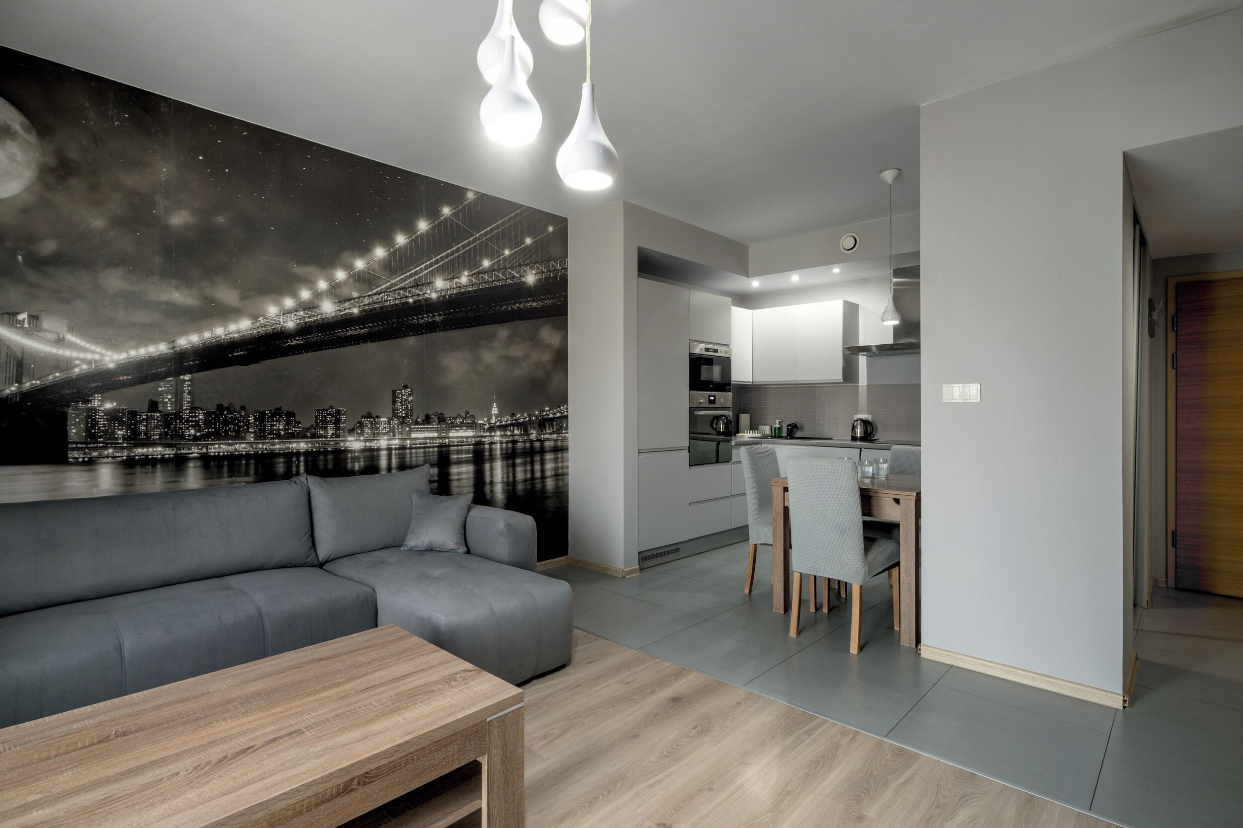 Un salon moderne avec un canapé gris, une table basse en bois et une fresque murale représentant un paysage urbain.