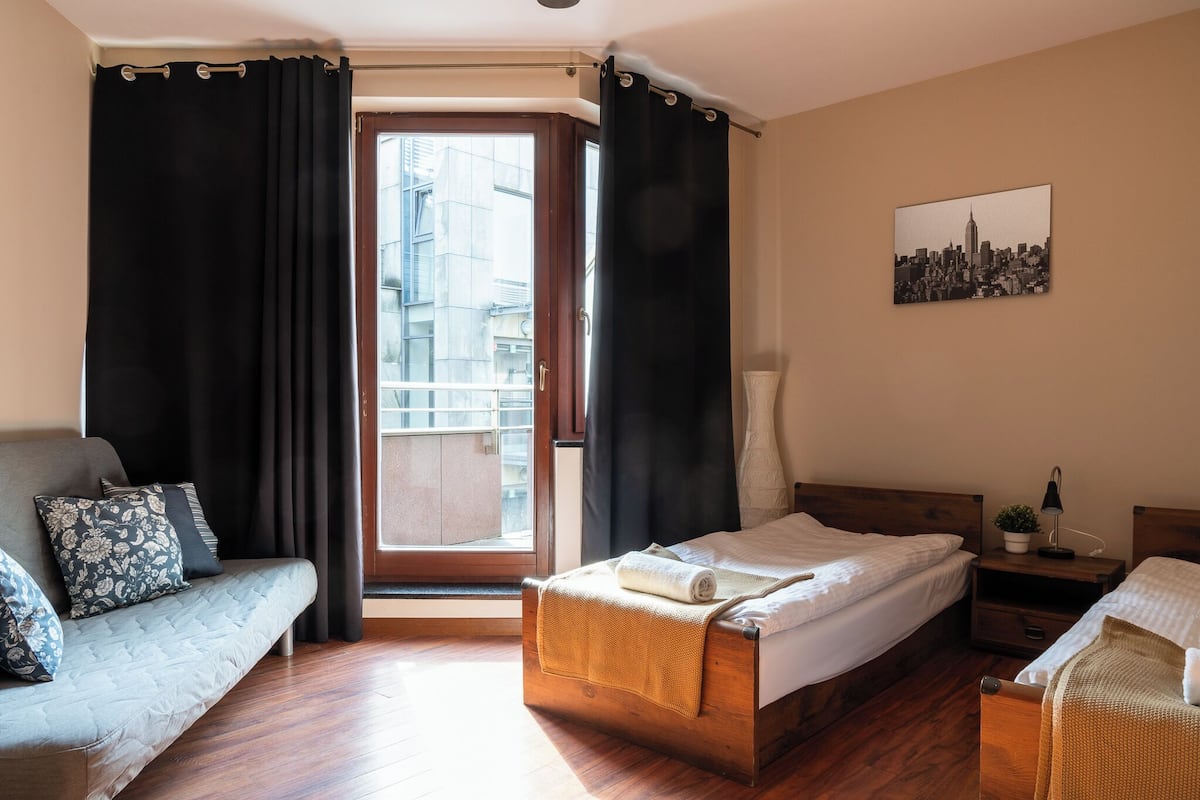 A hotel room with a bed, a sofa, a small table with a lamp, a window with curtains, and a cityscape picture on the wall.