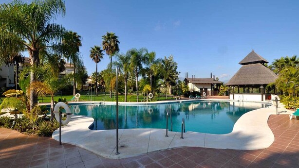 A swimming pool with a thatched-roof structure, surrounded by palm trees and a tiled deck. - El Campanario del Paraíso (Estepona)
