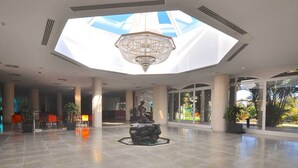 A spacious lobby with a large chandelier, a statue, and a view of greenery through large windows. - El Campanario del Paraíso (Estepona)