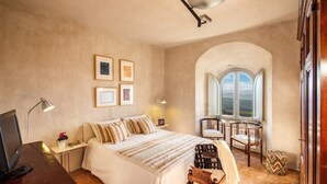 Ein Schlafzimmer mit Bett, Schreibtisch und einem kleinen Tisch mit Stühlen. Ein Fenster mit Bogenform bietet einen Blick ins Freie.