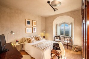A bedroom with a bed, desk, and a small table with chairs. There is a window with an arched design and a view of the outdoors.