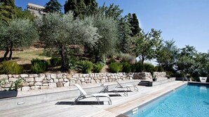 Ein Schwimmbecken mit Holzterrasse und Liegestühlen, umgeben von Grünpflanzen und einer Steinmauer.