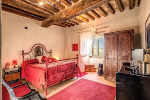 A bedroom with a wooden ceiling, a bed with a red bedspread, a nightstand with a lamp, a dresser, and a window with curtains.