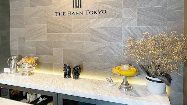 A reception area with a tiled wall displaying "THE BARN TOKYO" logo, a marble counter, and decorative items including a vase with dried flowers and a fruit bowl. - The Barn Tokyo (Tokyo)