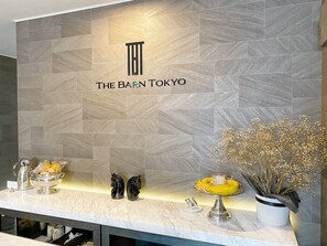 Un espace dâaccueil avec un mur recouvert de carreaux sur lequel est affichĂ© le logo « THE BARN TOKYO », un comptoir en marbre et des Ă©lĂ©ments dĂ©coratifs, notamment un vase avec des fleurs sĂ©chĂ©es et un panier de fruits.