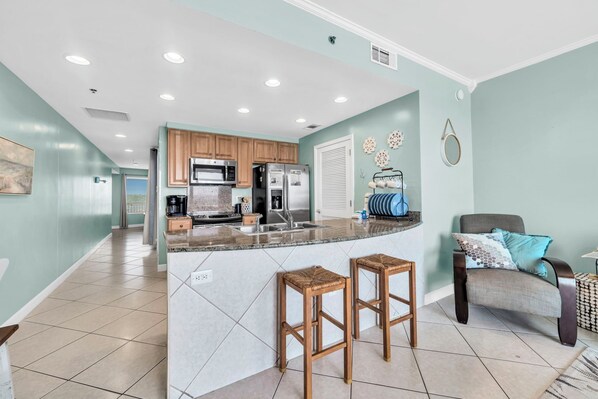 A modern kitchen with a central island, wooden bar stools, and a grey armchair with a cushion.