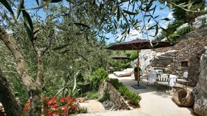 Patio esterno con pareti in pietra, ulivi e fiori rossi. Sono presenti due ombrelloni, un tavolo e sedie.