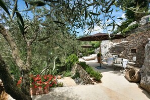 Eine Terrasse im Freien mit Steinmauern, Olivenbäumen und roten Blumen. Zwei Sonnenschirme, ein Tisch und Stühle sind ebenfalls vorhanden.