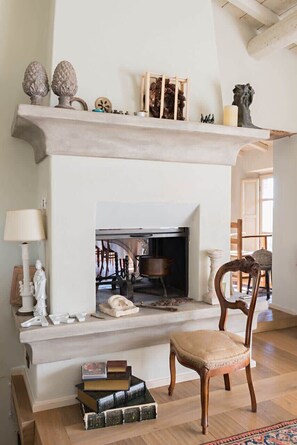 Una chimenea con repisas decoradas con diversos objetos, un sillón frente a ella, y una estantería llena de libros.