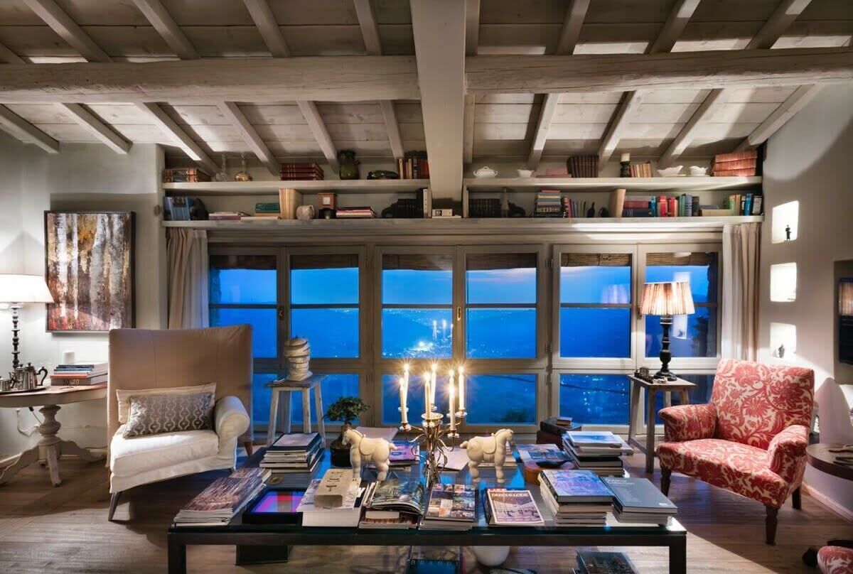 A cozy living room with a high ceiling, bookshelves, a coffee table with books, and a view through large windows.