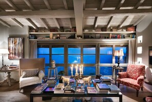 Un salon chaleureux avec un plafond haut, des étagères à livres, une table basse sur laquelle sont posés des livres, et une vue dégagée grâce à de grandes fenêtres.