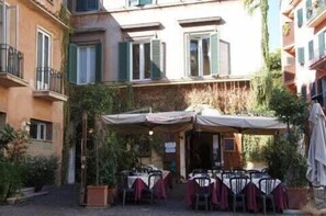 An outdoor cafe with tables and chairs, surrounded by buildings with balconies and shutters.