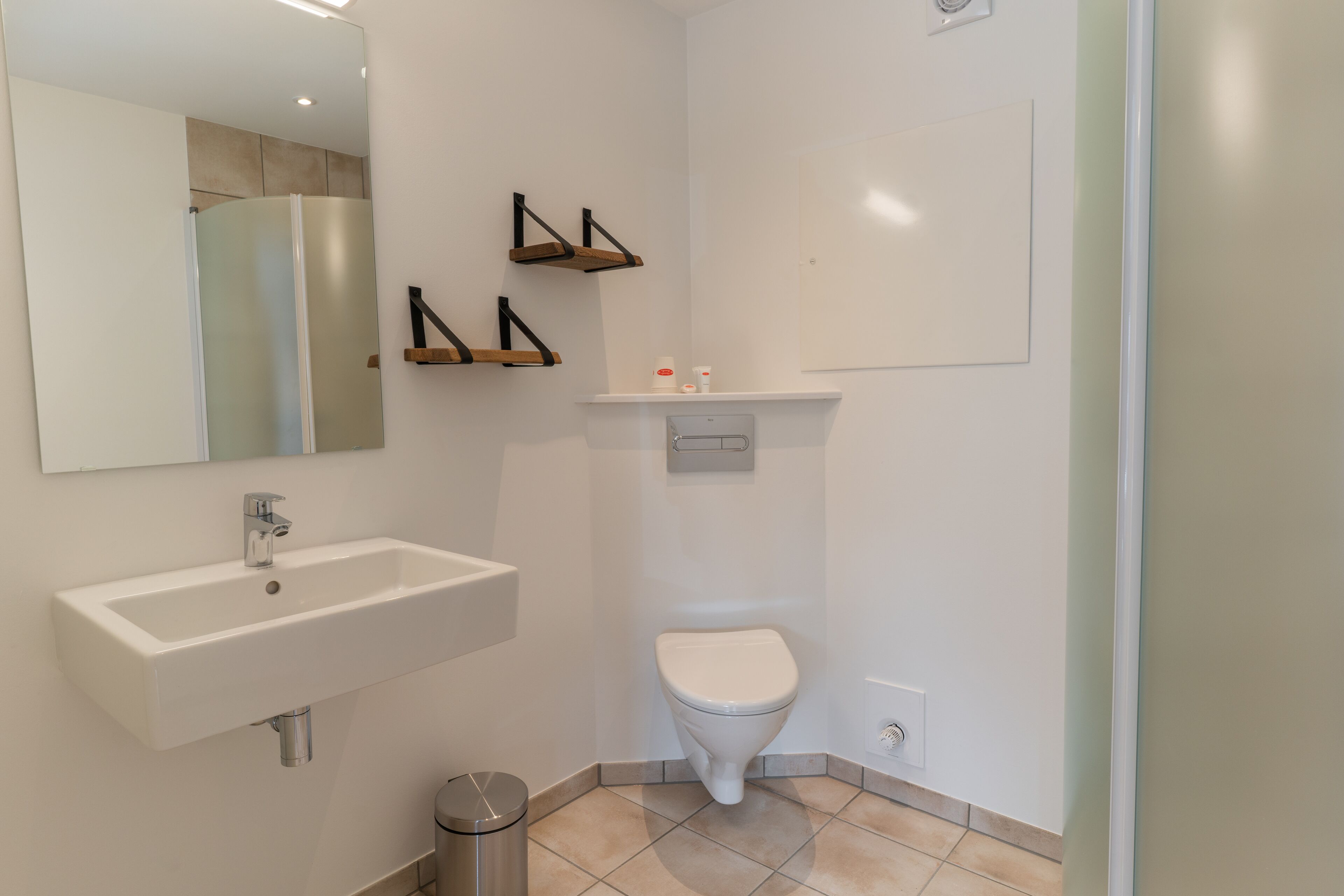 Ein modernes Badezimmer mit einem weißen Waschbecken, einer Toilette und einer Duschkabine. Über der Toilette befinden sich Holzregale und über dem Waschbecken ein Spiegel.