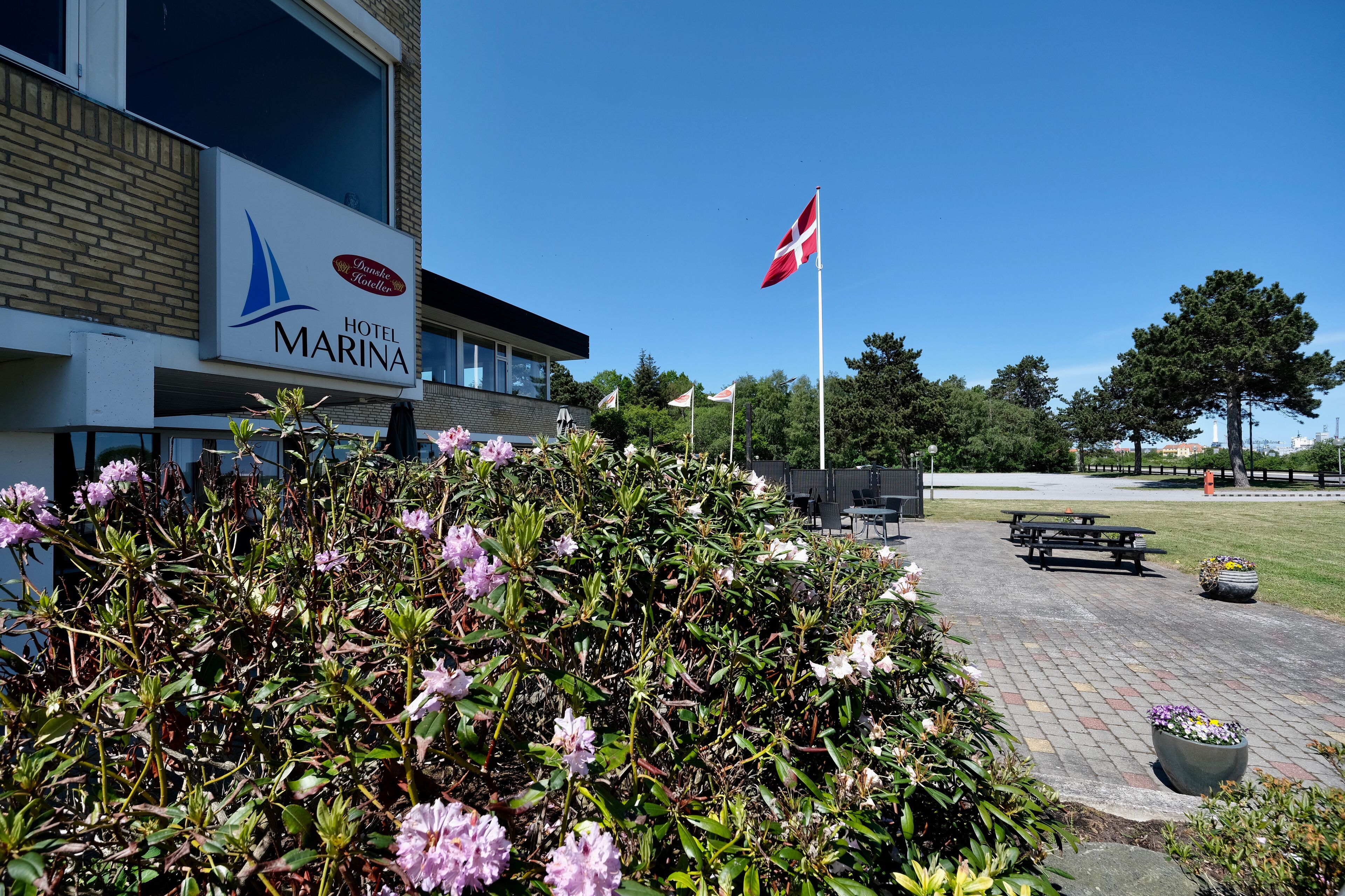 Un hotel llamado 'Hotel Marina' con vista despejada a la bandera danesa, un área exterior pavimentada con bancos y un jardín bien cuidado con flores rosas.