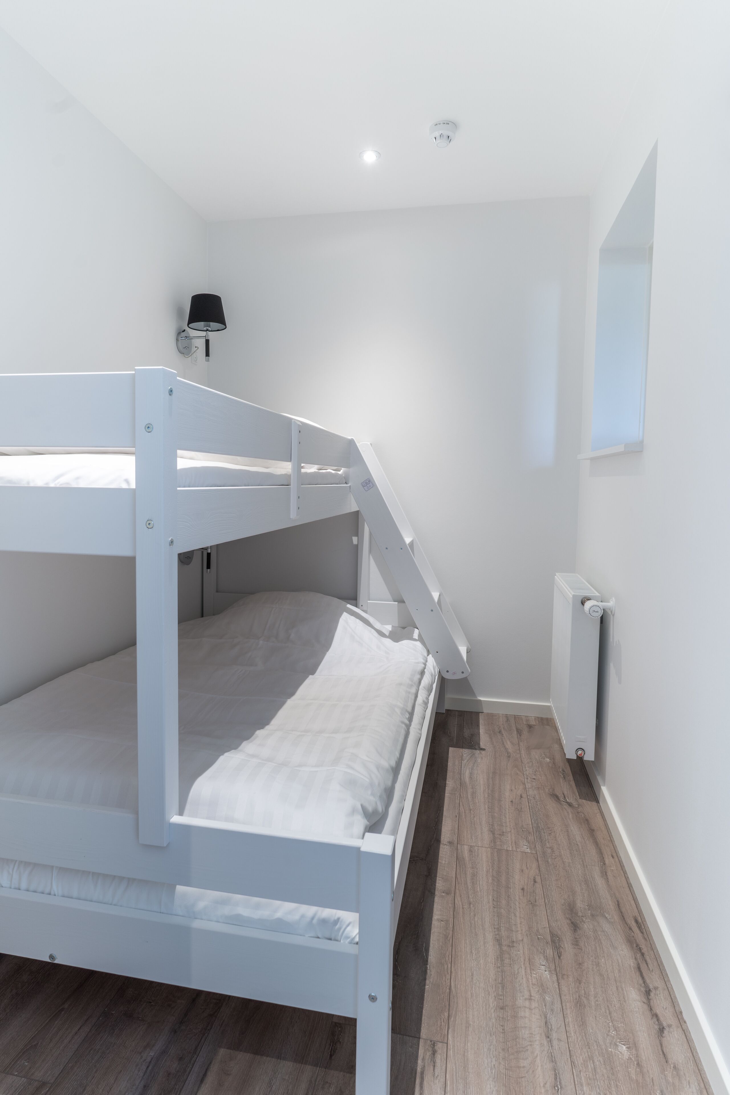 Habitación con literas, decoración en blanco, un radiador y una lámpara fijada a la pared.
