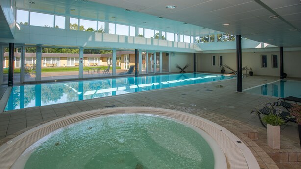 Una piscina interior con un área de jacuzzi separada, paredes de vidrio transparente y vistas al exterior.