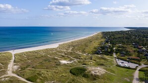 Vista aérea de una zona costera con una playa arenosa, dunas cubiertas de hierba y una hilera de casas a lo largo de la orilla.