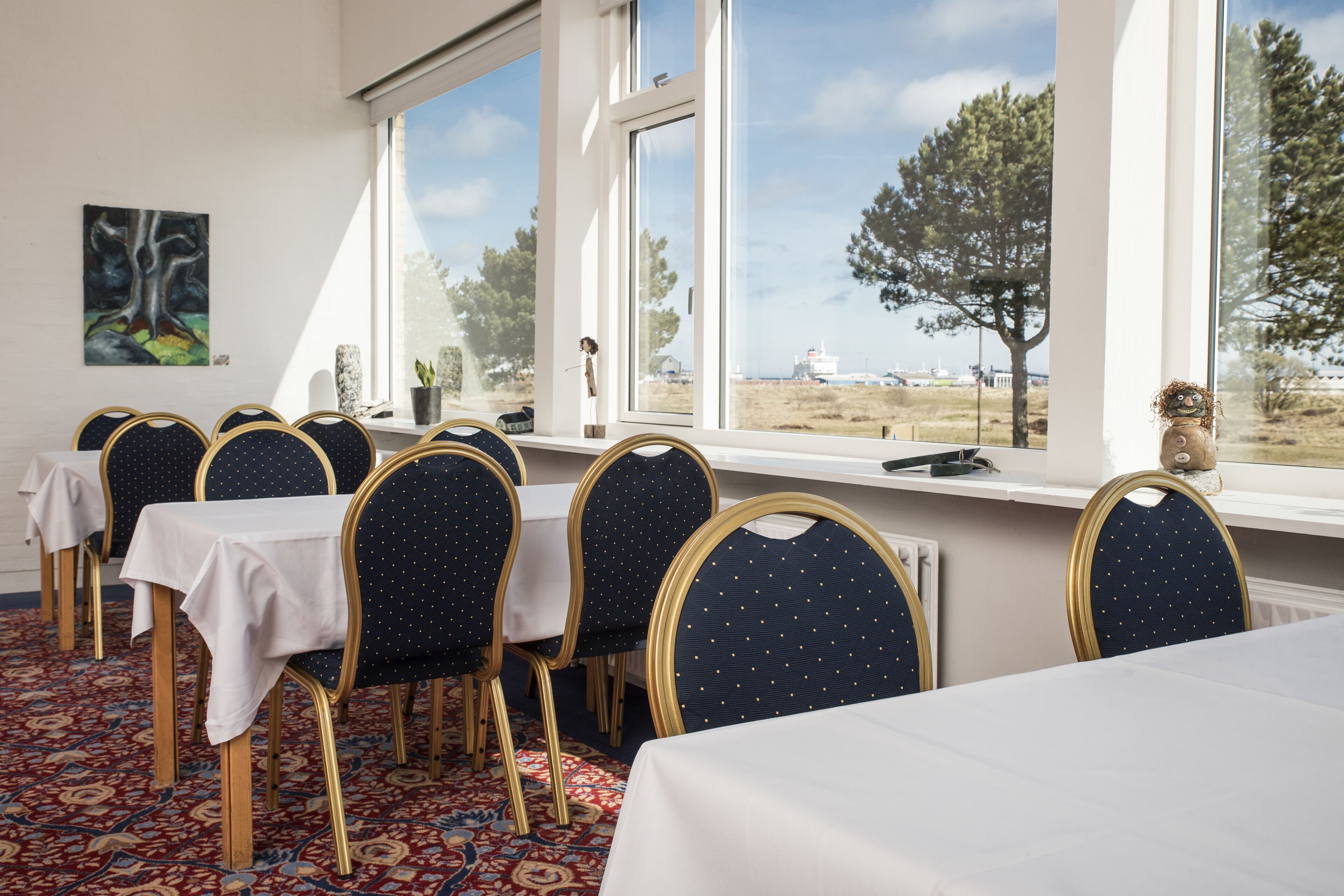 A dining area with tables and chairs, large windows with a view of the sea and trees, and a painting on the wall.