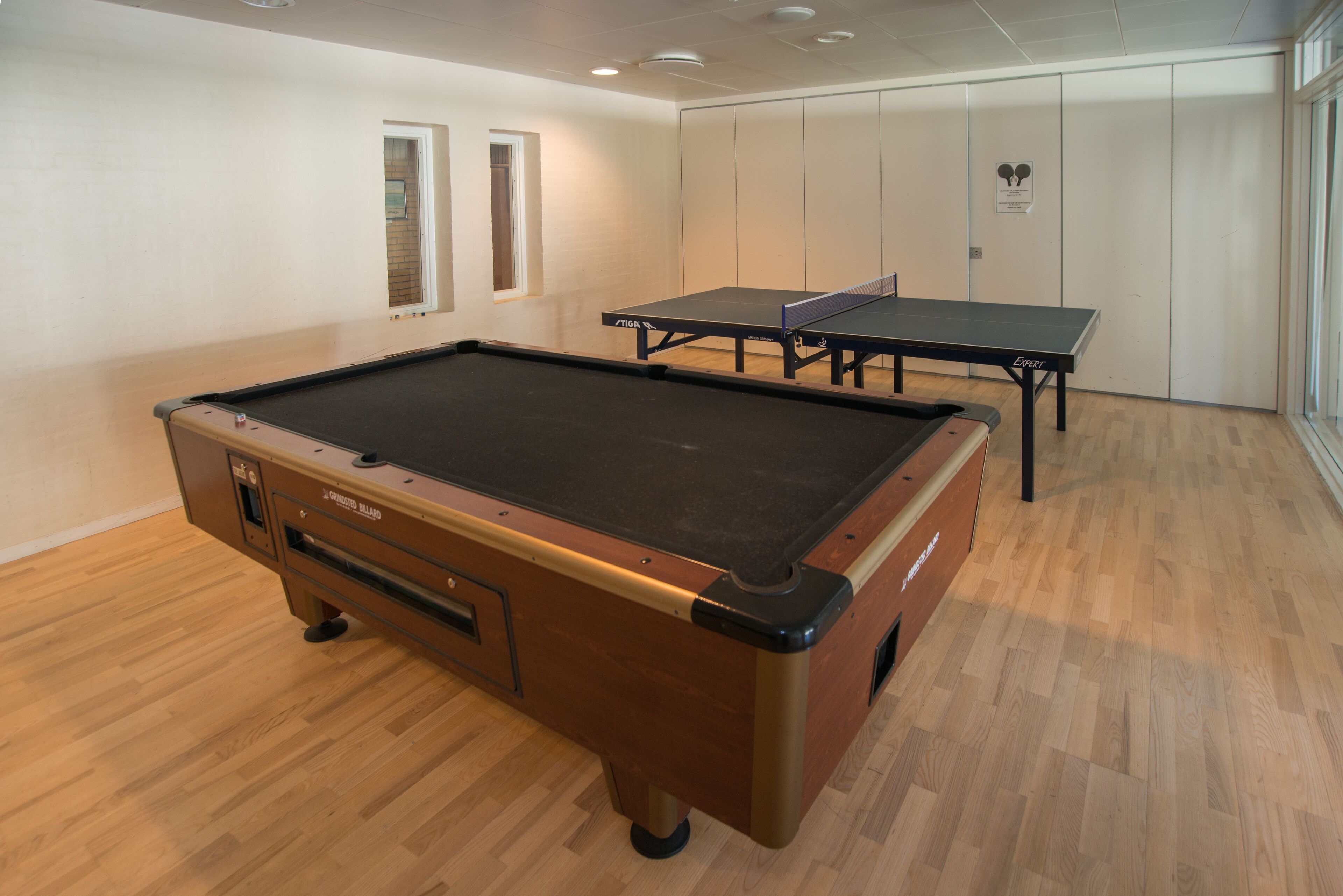 Une table de billard avec un sol en bois et une table de ping-pong dans une pièce aux murs blancs et dotée d’une porte.