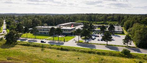 A hotel with a parking lot, surrounded by greenery and a road.