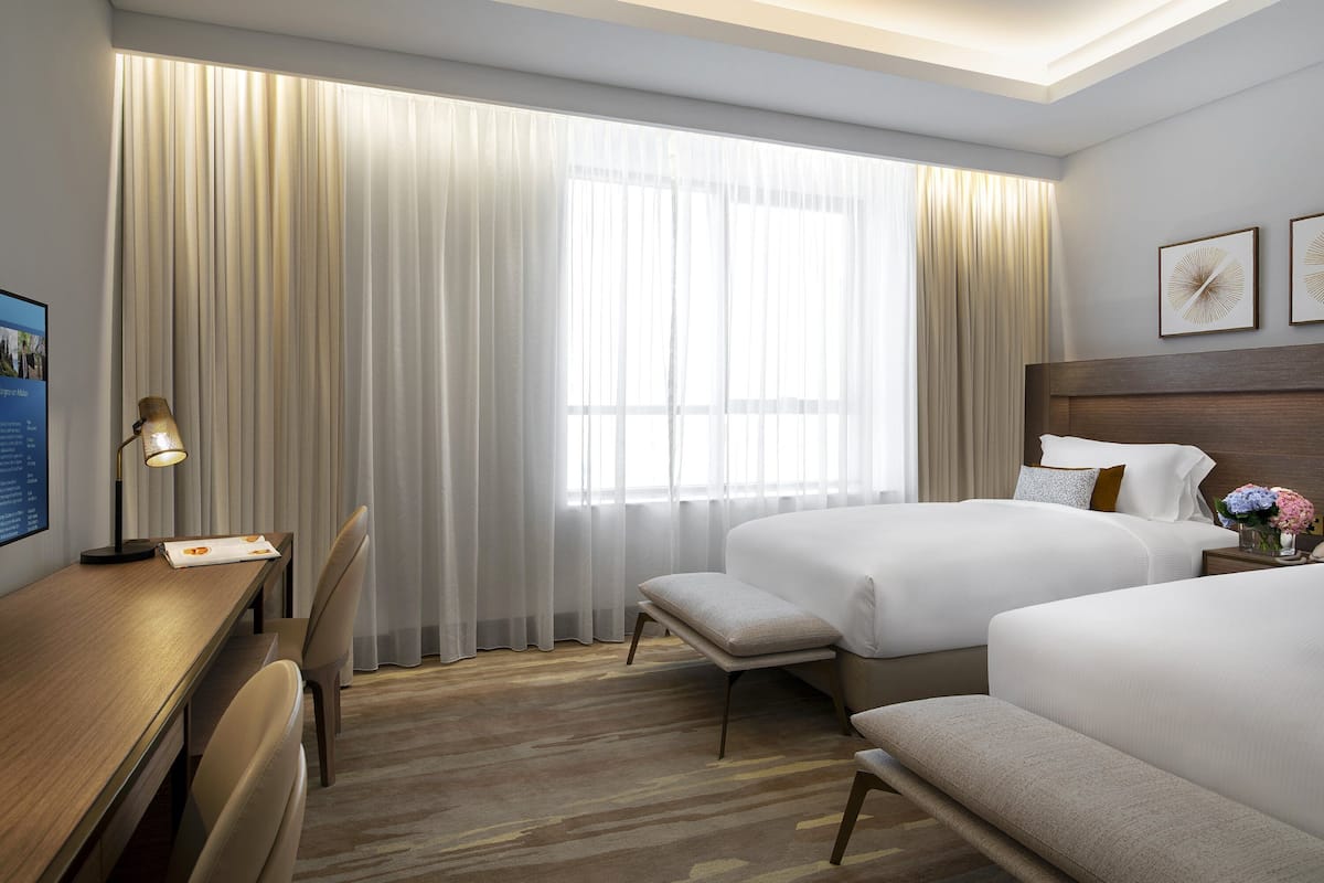 A hotel room with two beds, a desk, a chair, and a large window with curtains.
