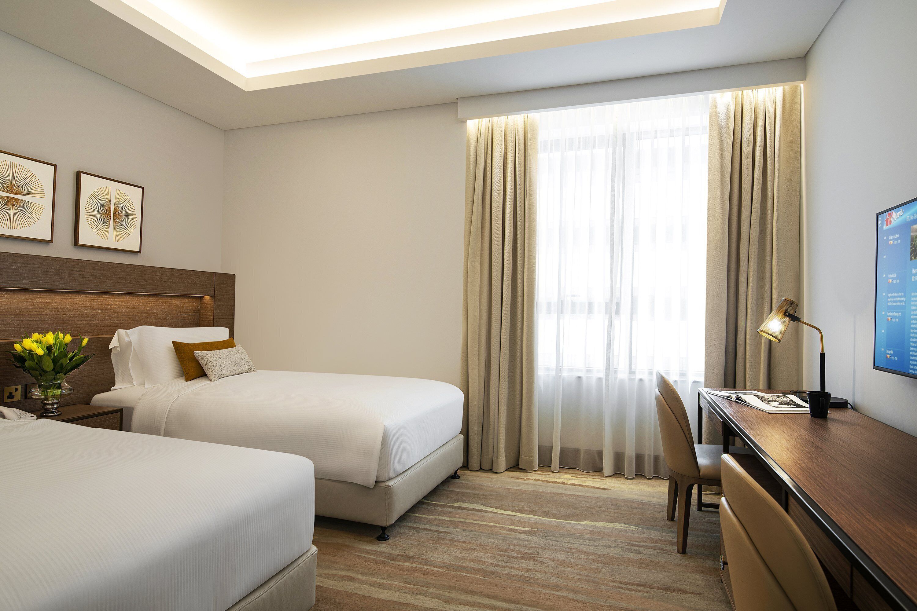 Une chambre d’hôtel avec deux lits, un bureau, une chaise, une lampe et une grande fenêtre avec des rideaux.