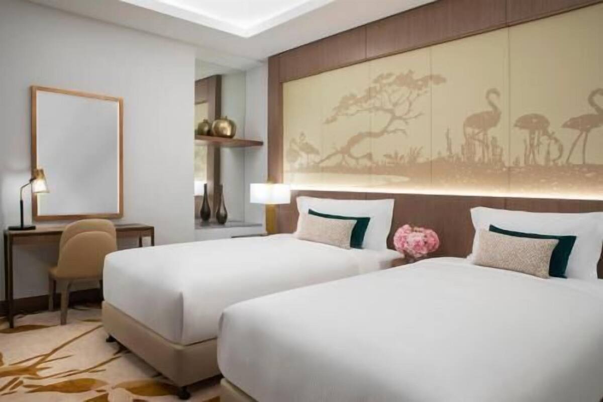 A hotel room with two beds, a desk, a chair, a lamp, and a wall with a tree and animal silhouette.