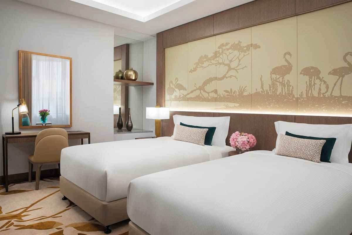 A hotel room with two beds, a desk, a chair, a lamp, and a decorative wall panel.