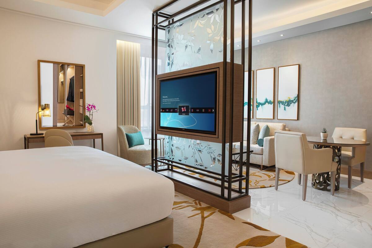 A modern hotel room with a large bed, a desk, a TV, and a seating area with chairs and a table.