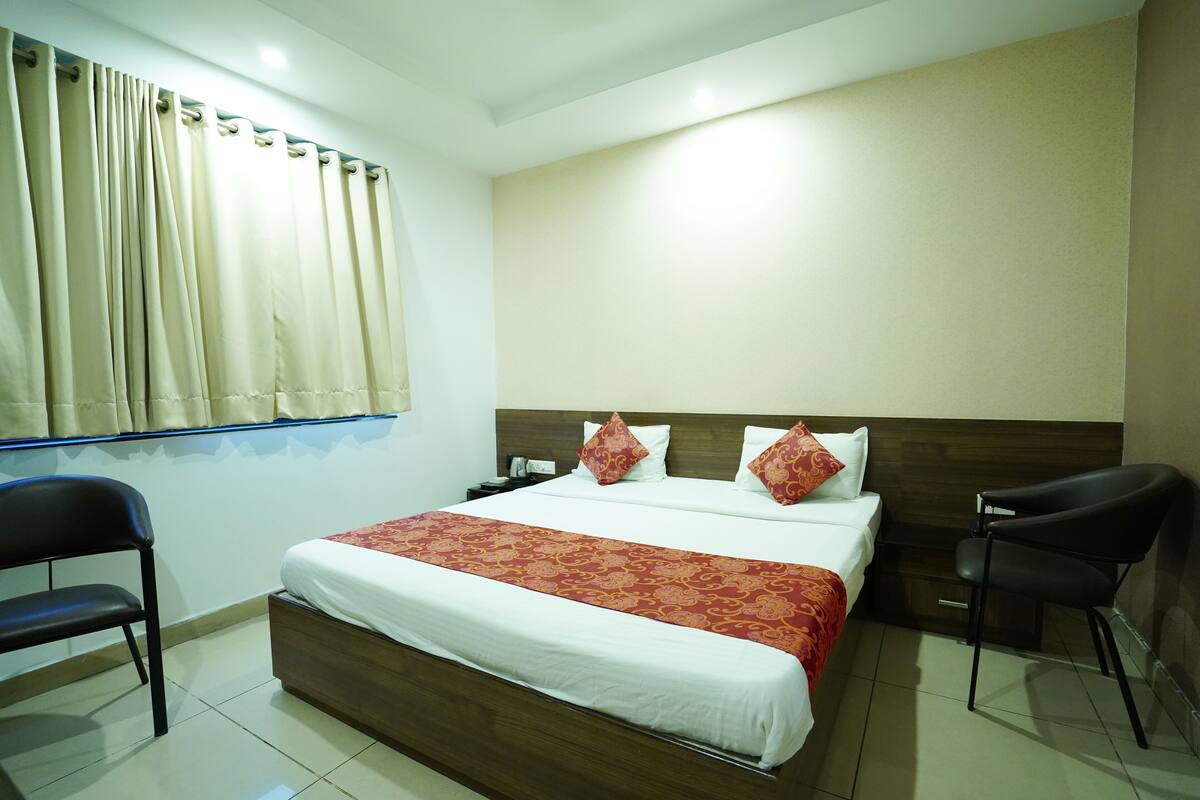 A hotel room with a large bed, two nightstands, a wooden headboard, a window with curtains, and two chairs.