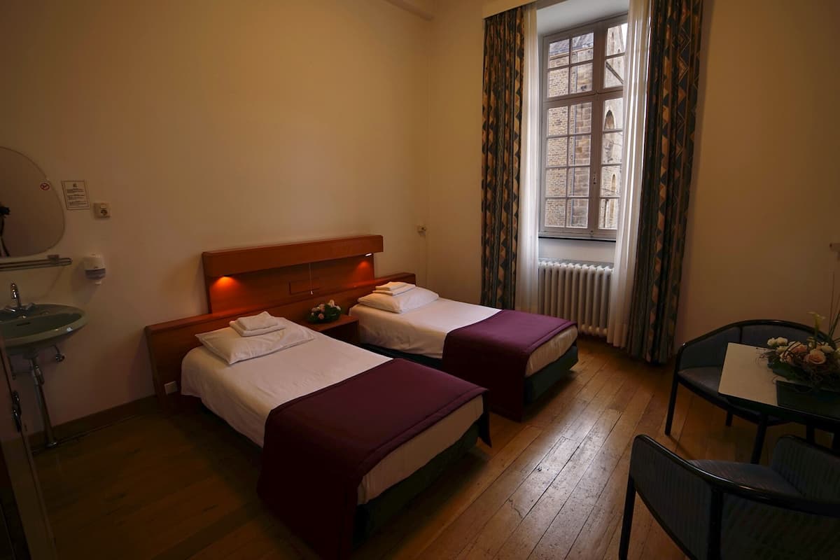 A hotel room with two beds, a small table with a chair, a window with curtains, and a radiator.