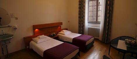 Una habitación de hotel con dos camas, una mesa pequeña con una silla, una ventana con cortinas y un radiador.