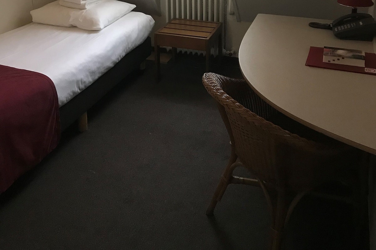 A compact hotel room with a bed, a small table, a chair, a radiator, and a window with curtains.